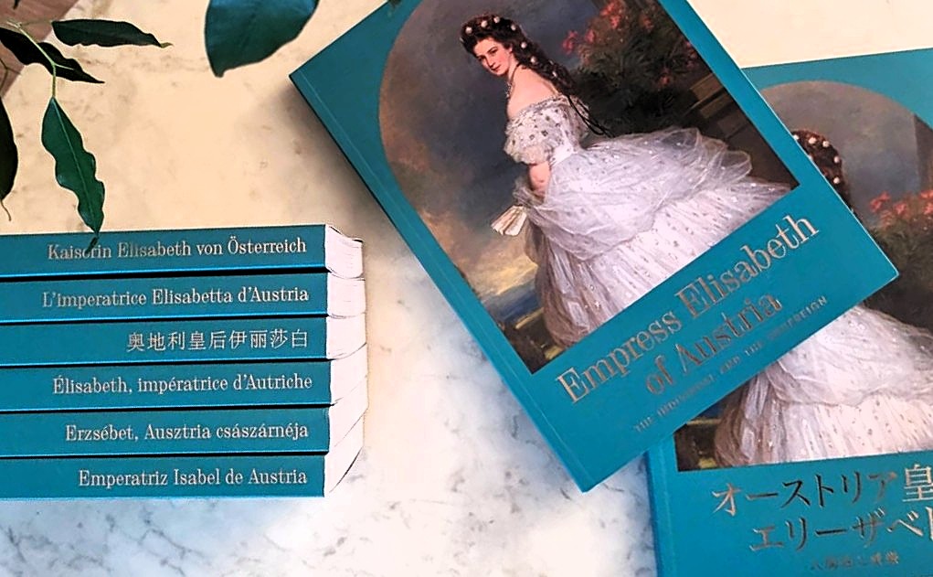 Sisi Biographie Übersetzungen Sisi Biographie in allen sieben Sprachen auf einem Tisch mit dekorativen Blättern.